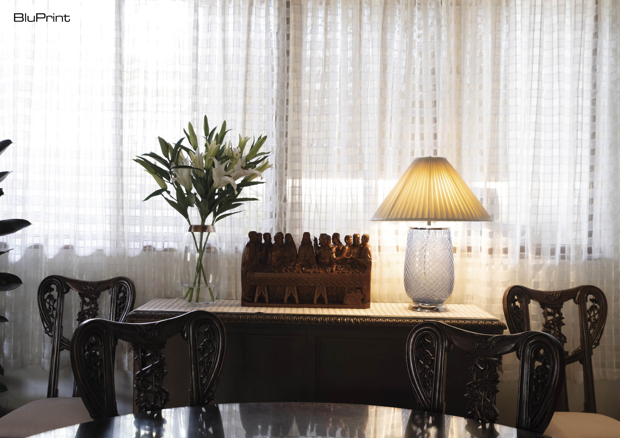 A traditional sideboard with a table lamp, floral arrangement, and wooden art piece next to a window with sheer curtains
