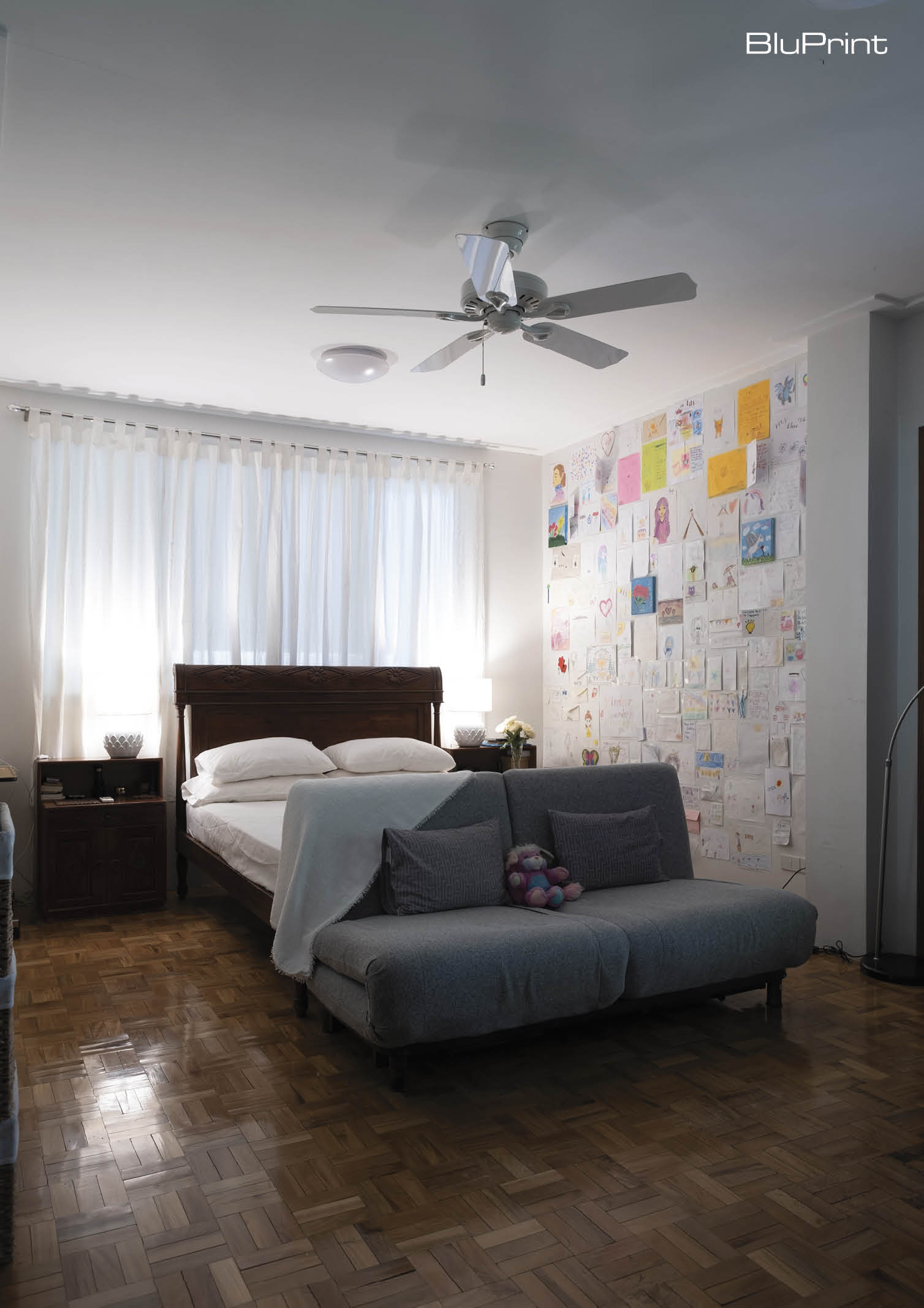 A kid's bedroom with a wooden bed frame and side tables. A two-seater couch is at the foot of the bed, and a wall displays the child's art work