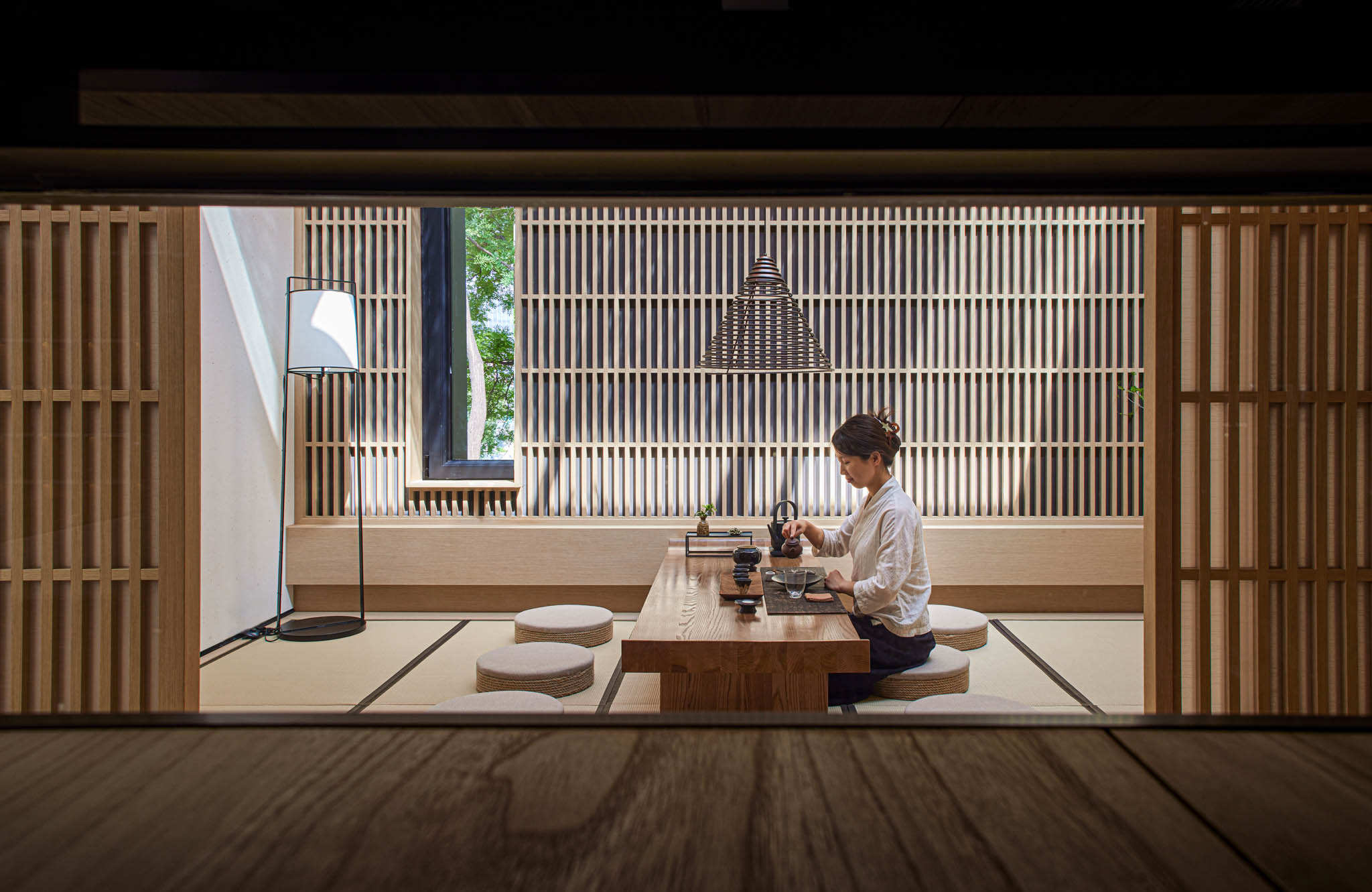 a person sits in a japanese tatami room with a low table and round cushions surrounded by bamboo screens that overlook the outside.