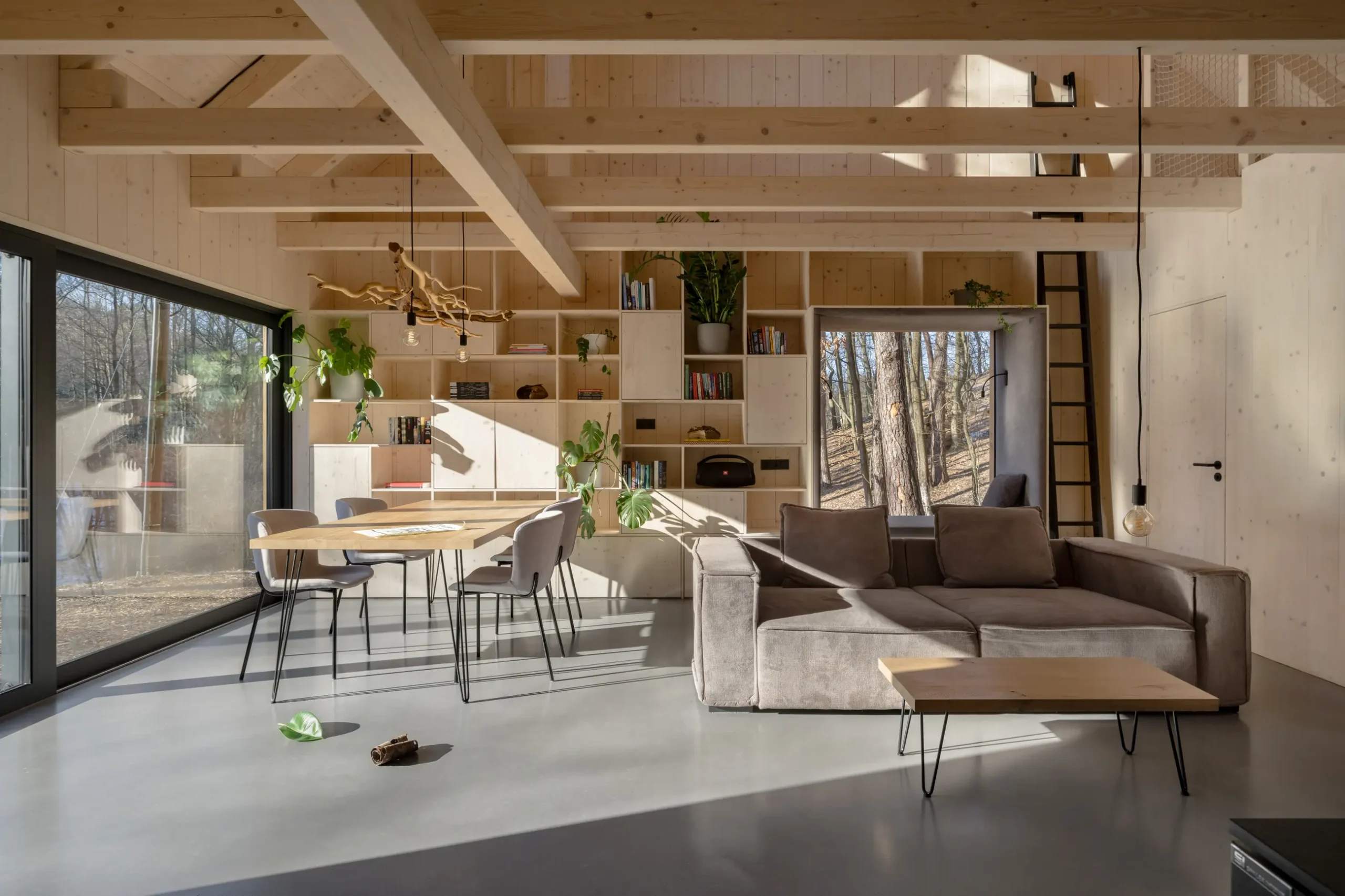 Living area of a wooden cabin with concrete floors, fabric couch, and dining area with wooden tables and chairs next to open shelving.