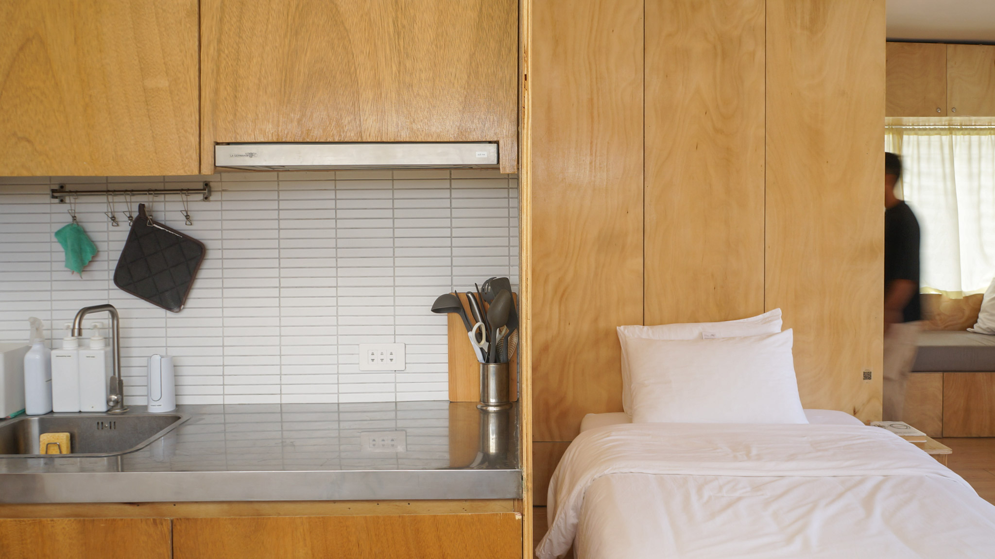 A small kitchenette with plywood joinery next to a double bed with white linens.