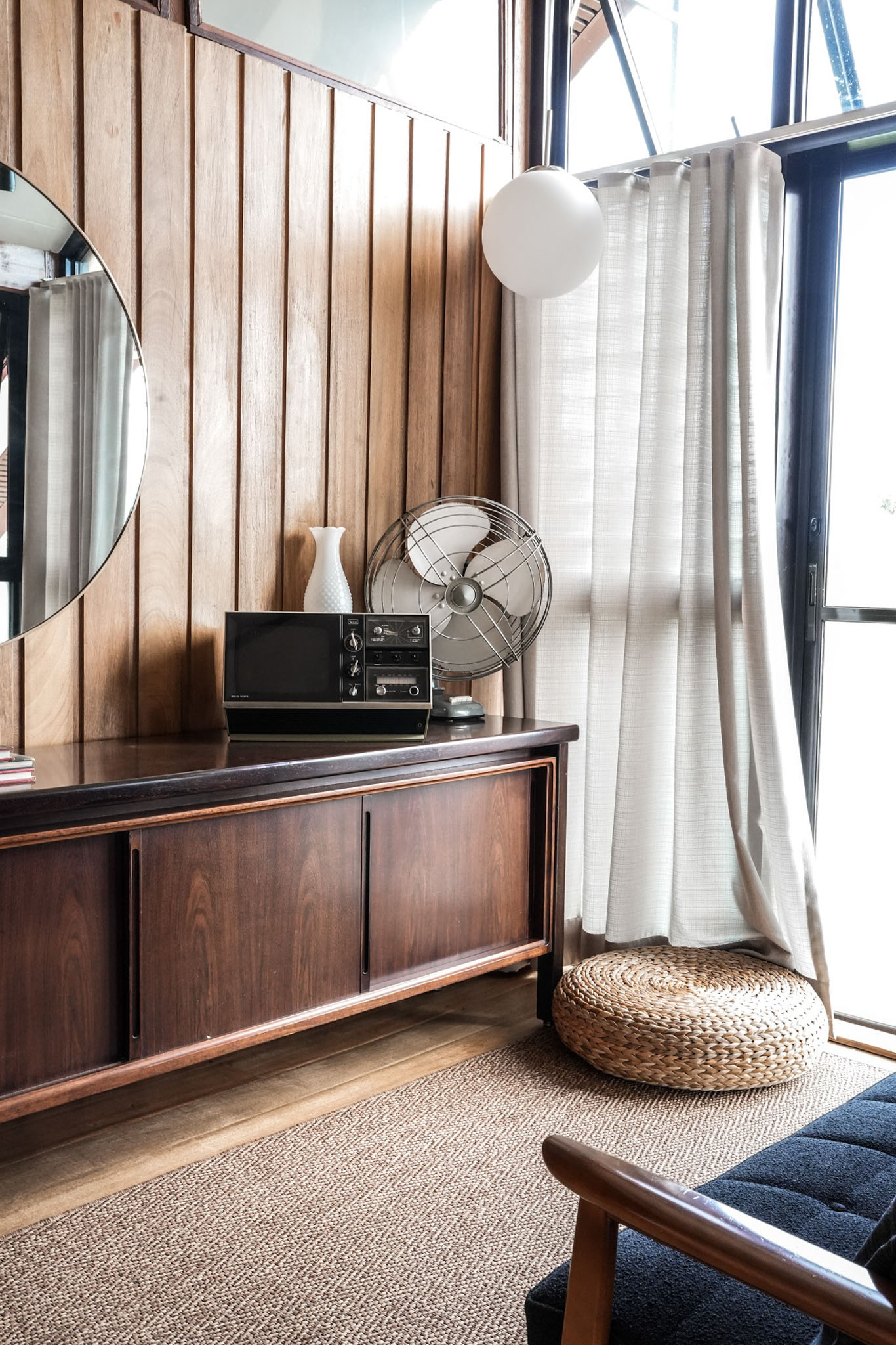 A room with a wooden console, round woven pouf, and an old electric fan. There is a brown woven rug and wooden wall panels.