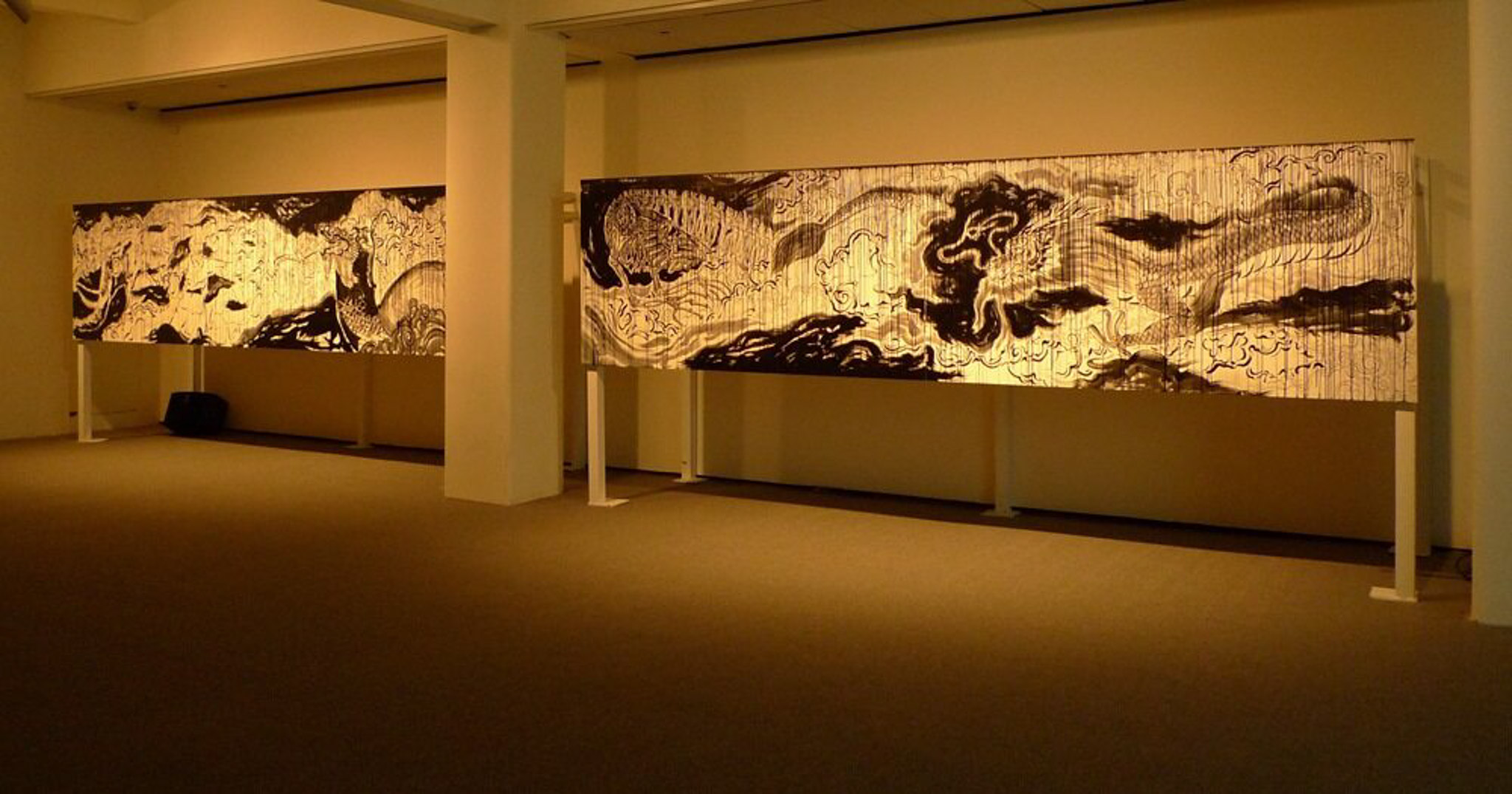 An abstract mixed media work of art mounted on a wall illuminated by a light yellow hue.