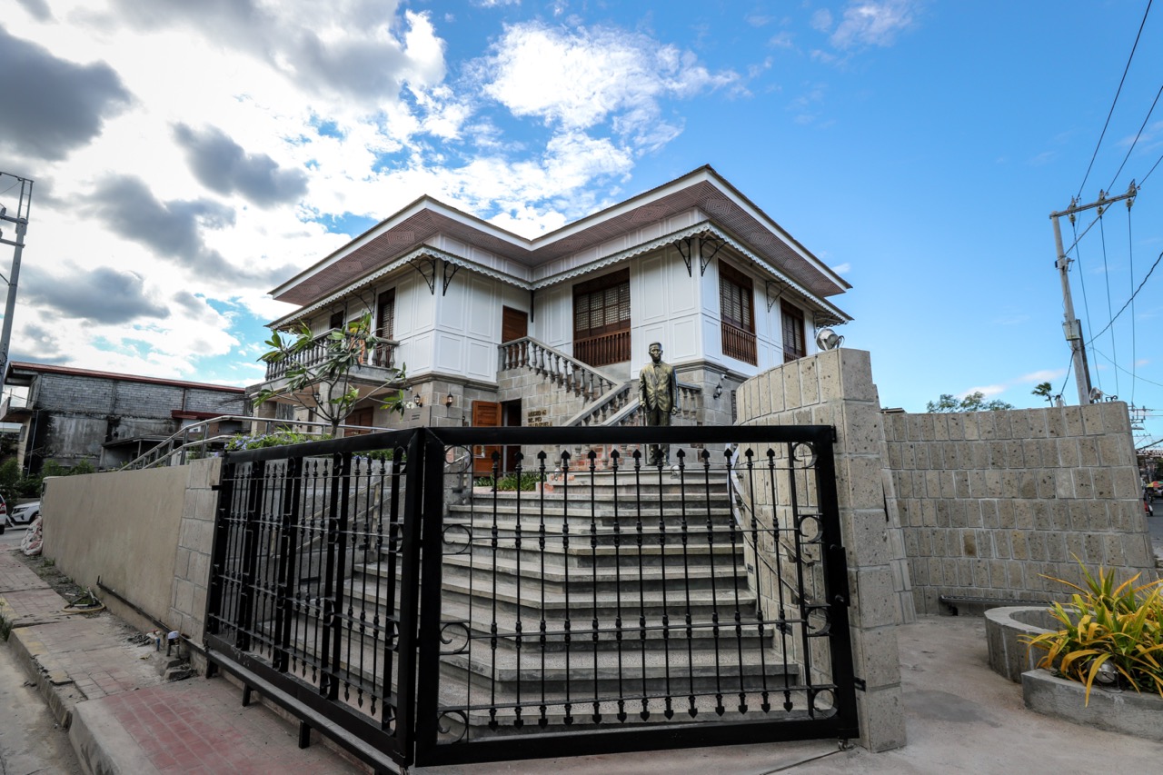 A view of Dr. Pio Valenzuela's bahay na bato, now a museum, with a metal gate and stone steps leading up to his statue.