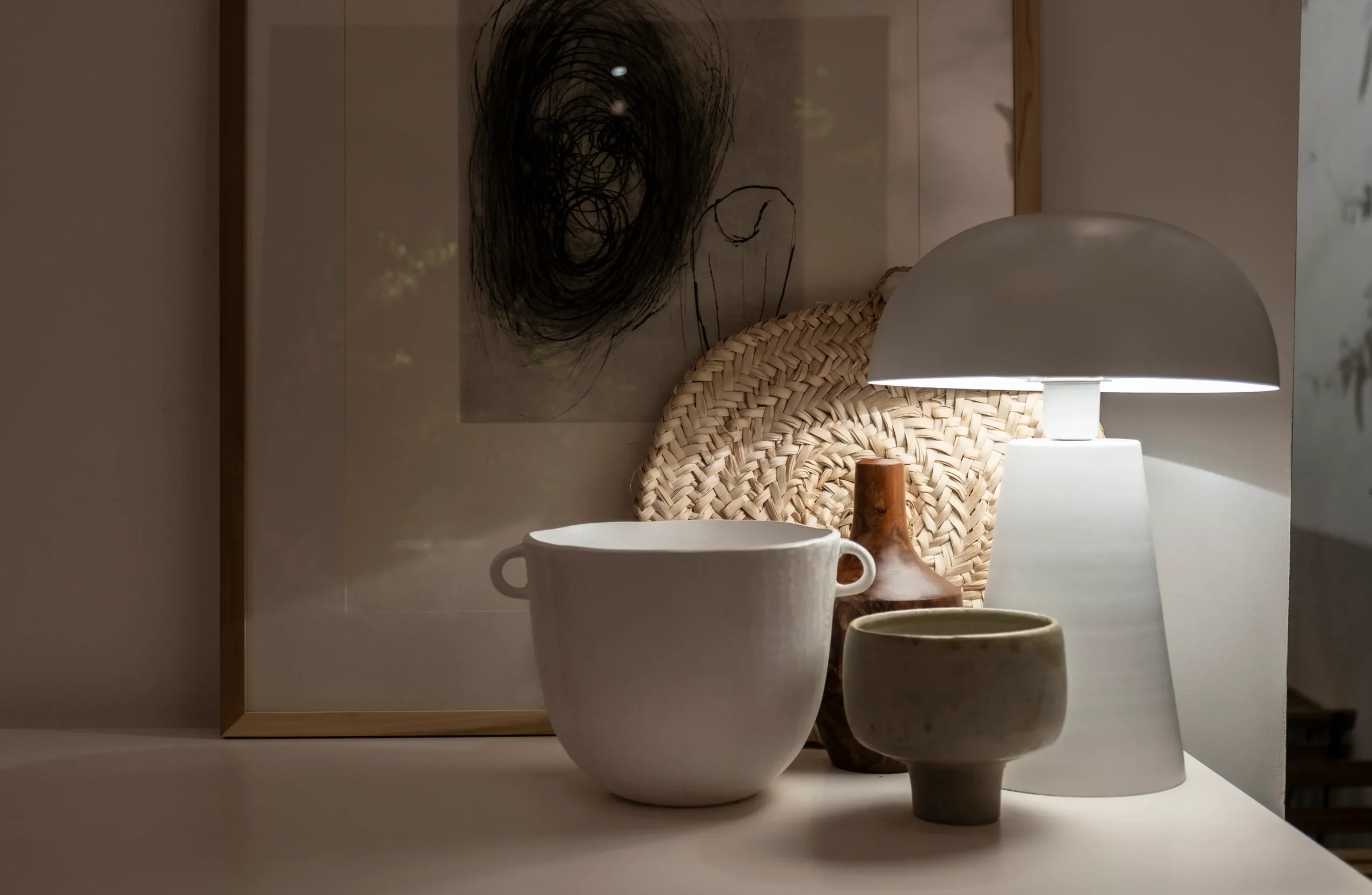 White table lamp illuminating ceramic vessels beside a woven tray and framed artwork on a neutral console.