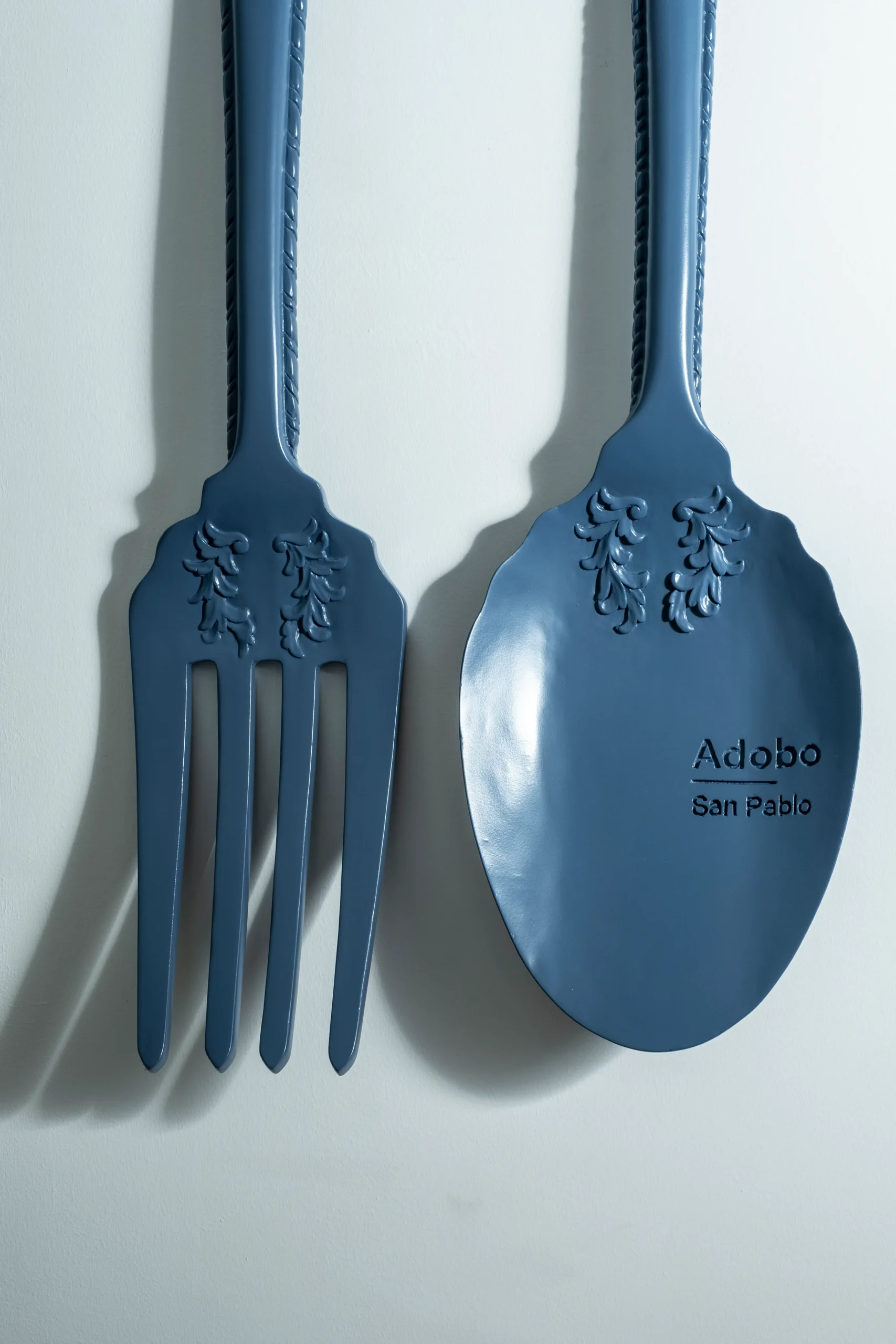 Oversized blue Kubyertos wall-mounted spoon and fork by Ito Kish engraved “Adobo San Pablo.”