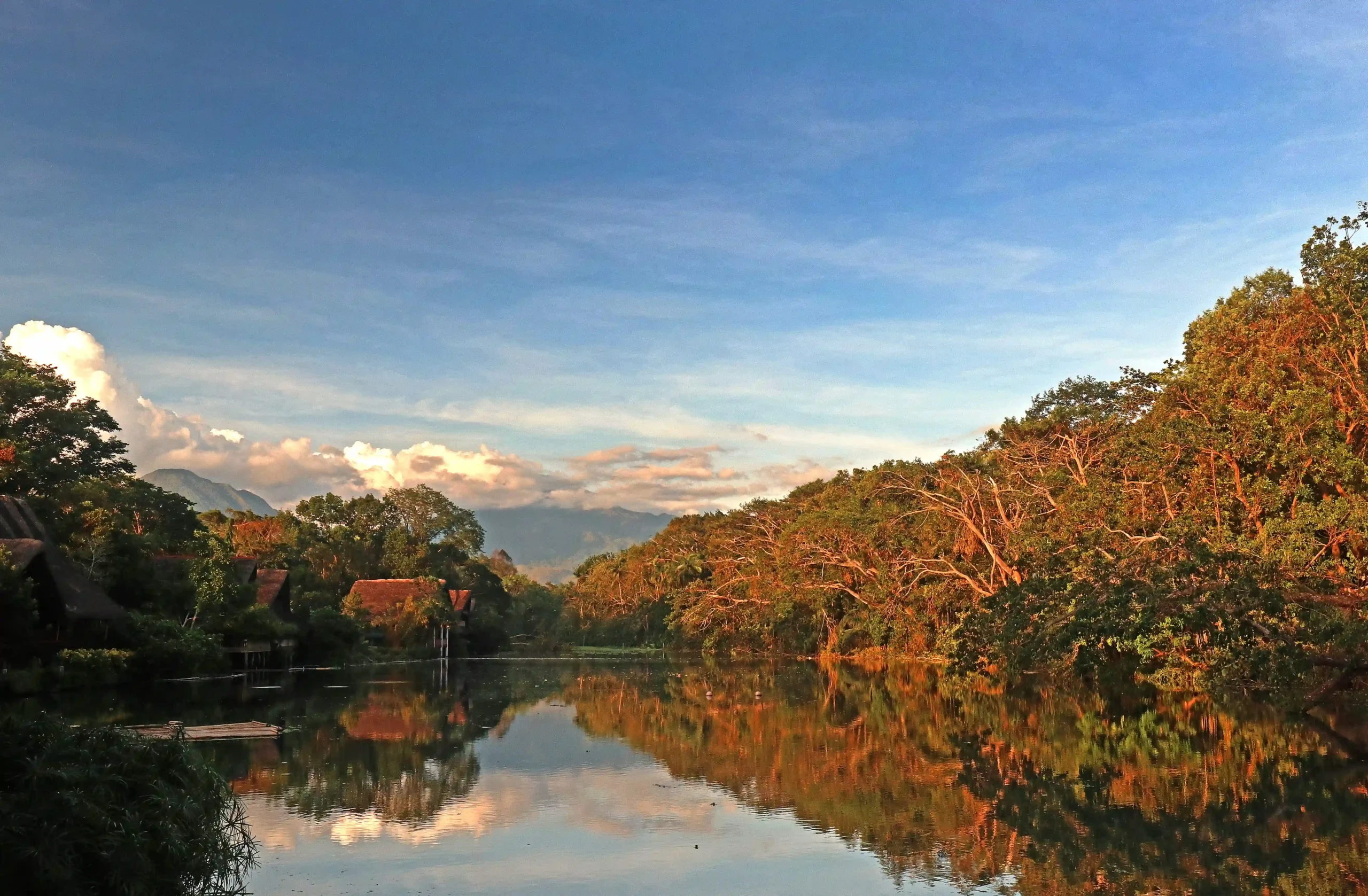 Sunlit river reflecting a blue sky and surrounding trees, with distant hills and a few houses on the left.  Villa Escudero