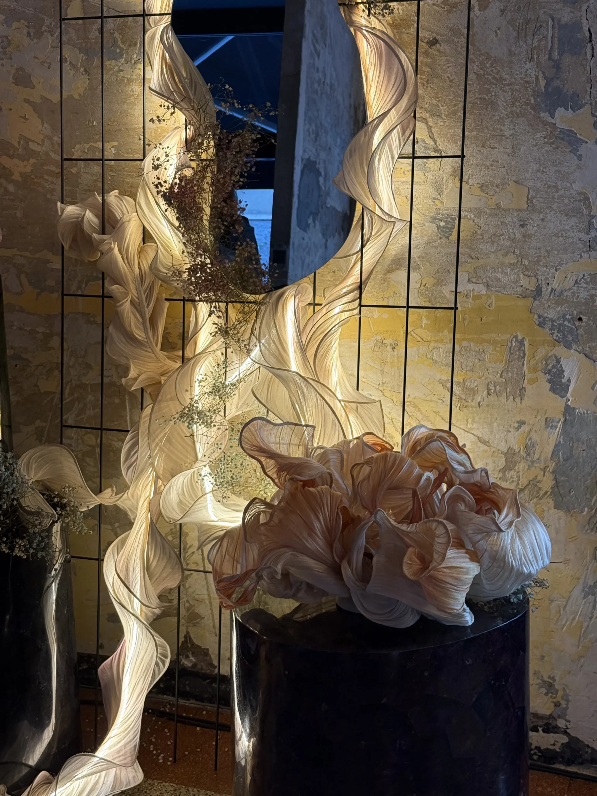 Art installation with flowing cream-colored fabric ribbons wrapped around a tall central column, set against a weathered wall.