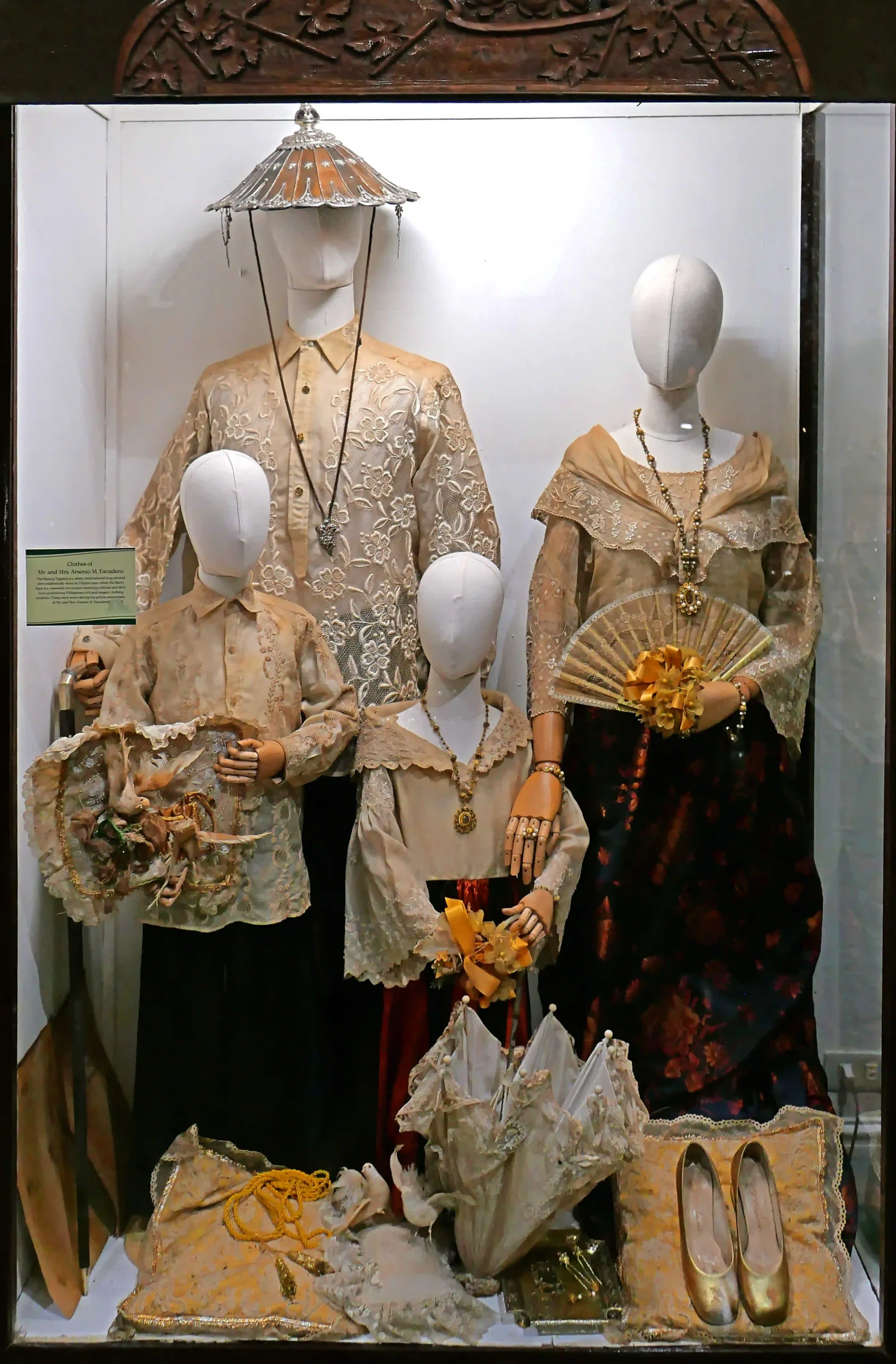 Window display of beige lace dresses on mannequins, with a decorative parasol hat, jewelry, and gold flats. The outfits are ornate and vintage-inspired; accessories fill the lower foreground.  Villa Escudero Plantation