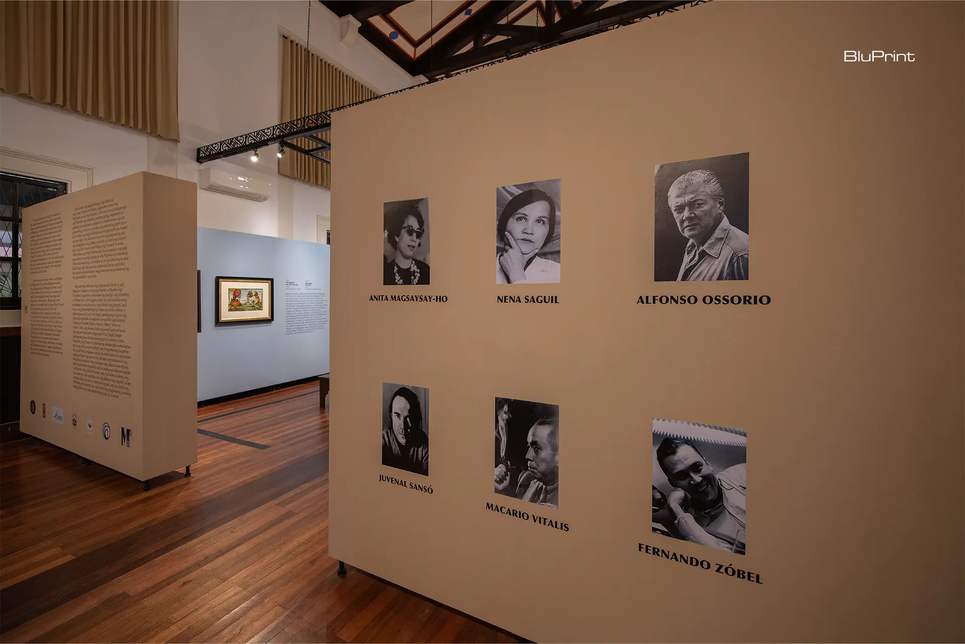 Exhibition wall with black-and-white portraits and names of artists like Anita Magsaysay-Ho, Nena Saguil, Alfonso Ossorio on beige panels; BluPrint logo top-right.
