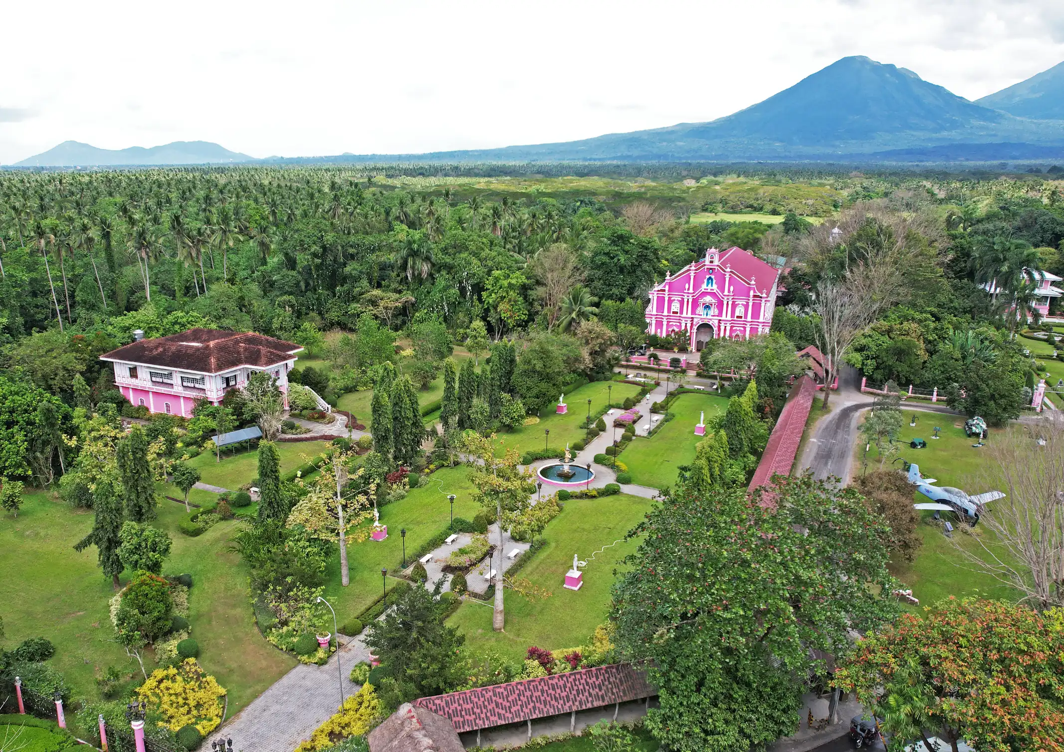 Aerial view of a pink church surrounded by lush gardens and tall palm trees, with blue mountains in the distance.
Villa Escudero