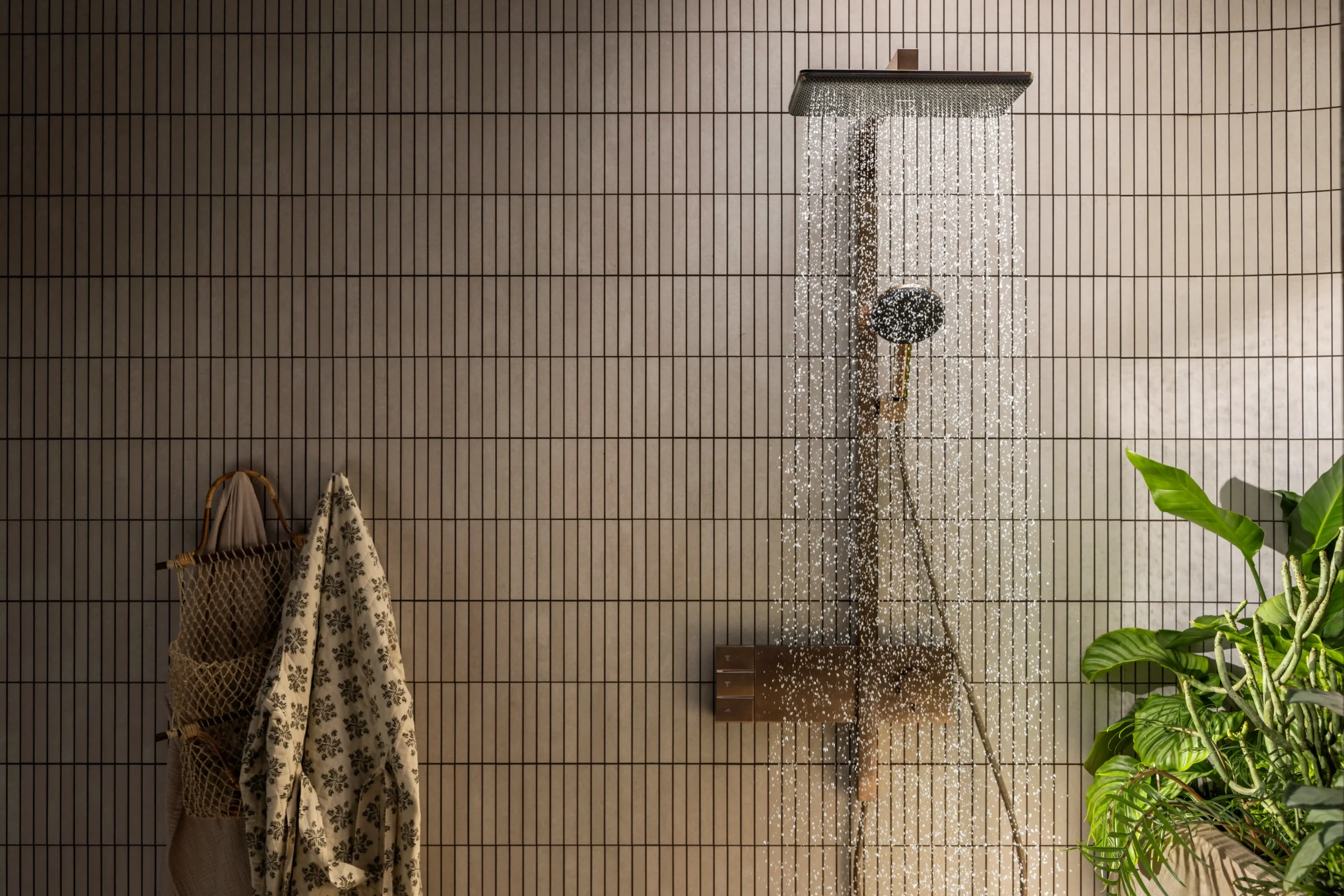 Beige tiled bathroom wall with a rainfall showerhead spraying water; a towel and woven bag hang on a rack to the left, and a green plant sits to the right.