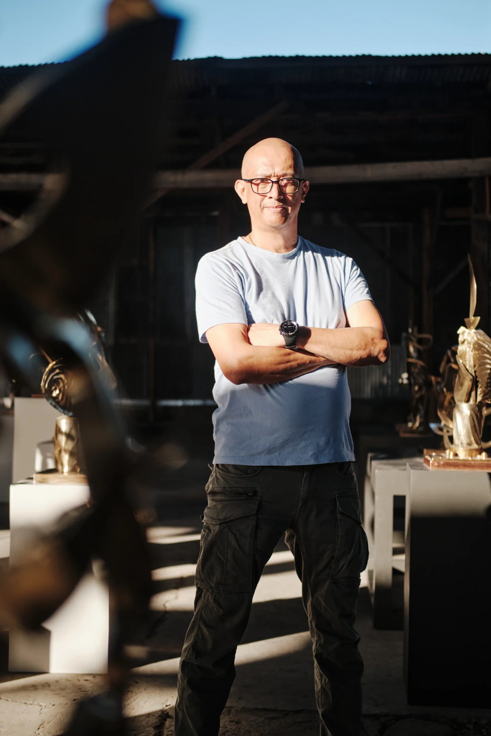 Bald man with glasses stands with arms crossed in a sunlit workshop, trophies visible in the background.  Anton V. Quisumbing, Sculpture, Pasulong