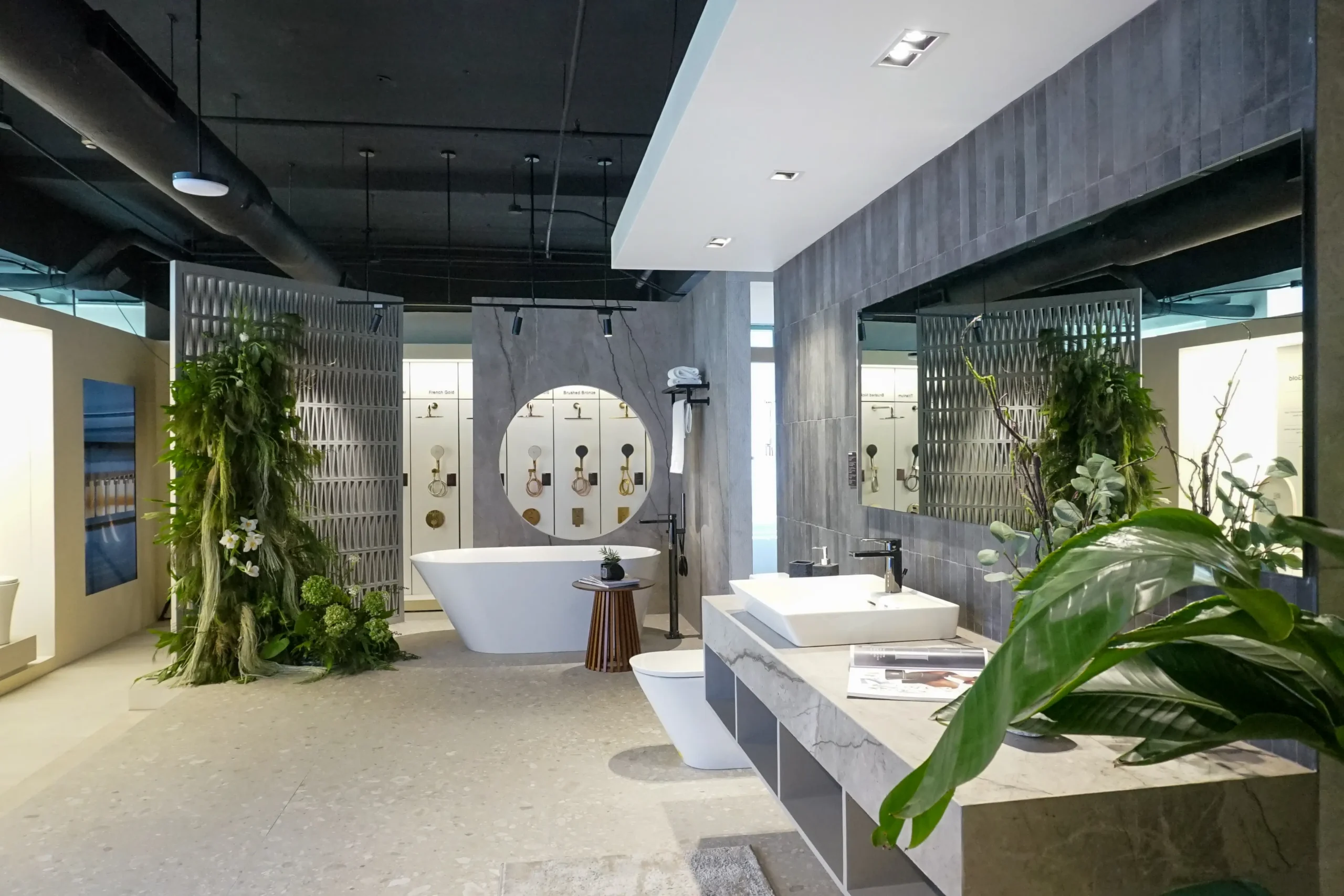 Modern bathroom showroom with a curved white freestanding tub, marble vanity, and a large round mirror; fixtures displayed on a gray wall behind.
