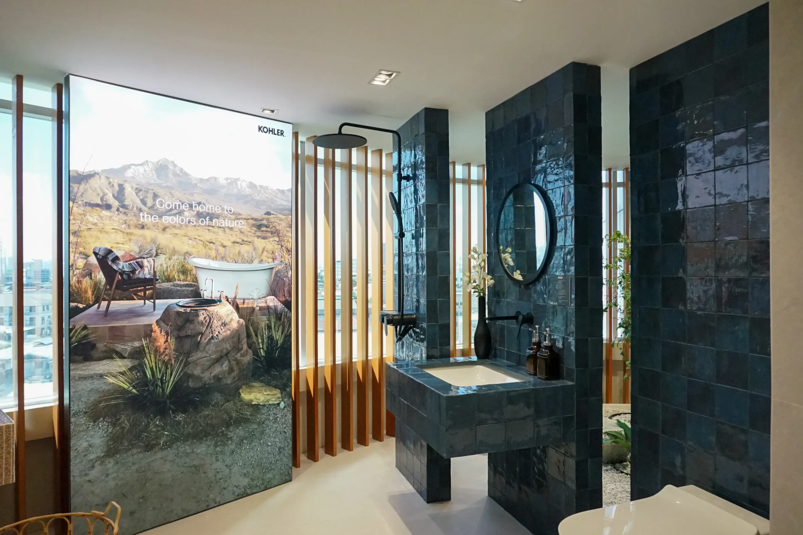 Showroom bathroom with dark tiled walls, a circular mirror, and a freestanding bathtub; promotional wall reads 'Come home to the colors of nature' with a mountain landscape.