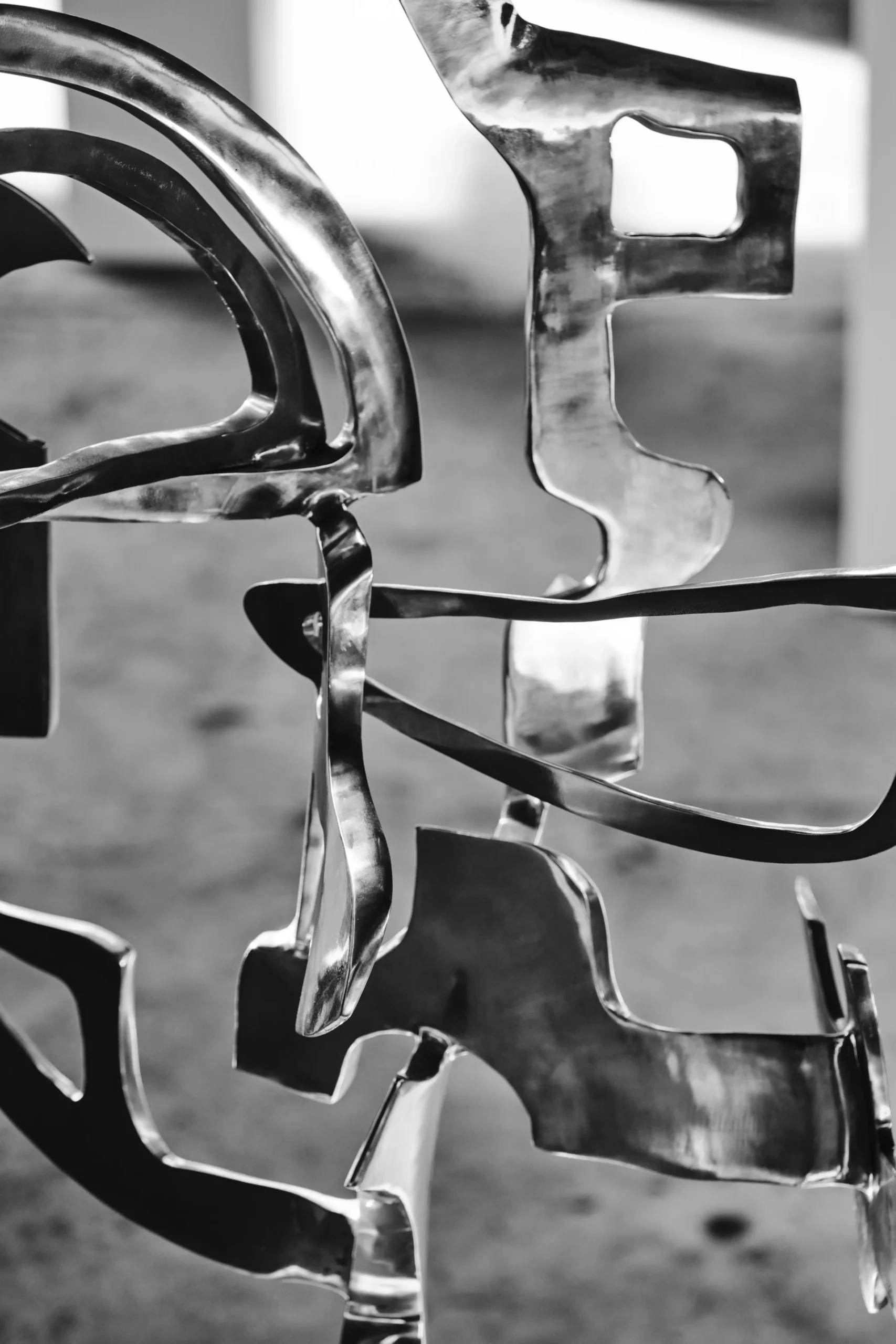 Close-up of abstract metallic ribbons twisting and curving, forming a chrome sculpture against a blurred background, in black and white  Anton V. Quisumbing, Sculpture, Pasulong