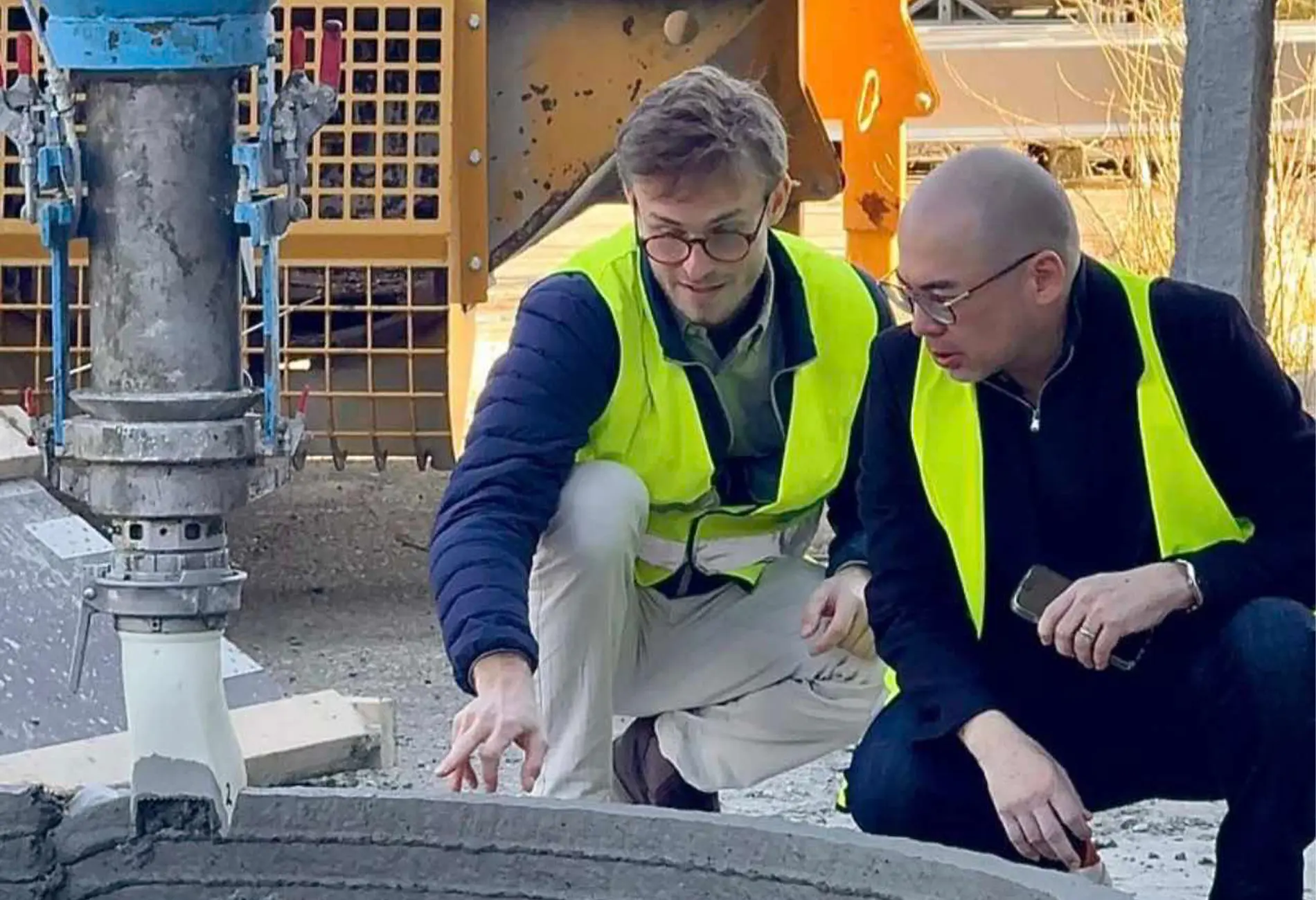 Two construction workers in yellow high-visibility vests kneel and inspect a concrete slab on a building site, with heavy machinery in the background.